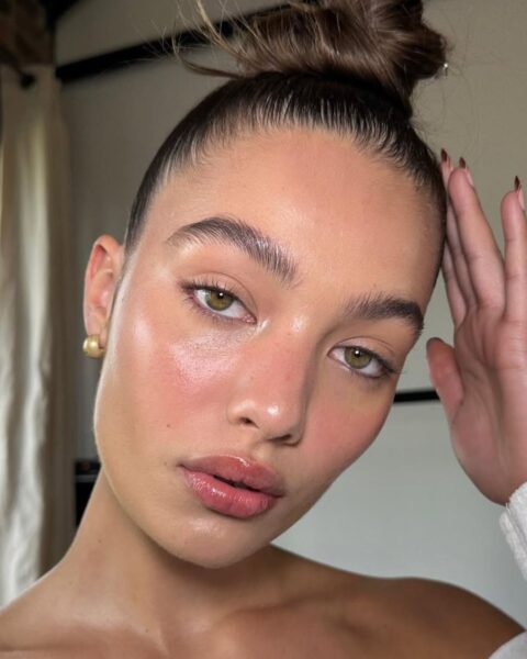 Zbliżenie na twarz kobiety z włosami upiętymi w gładki, wysoki kok i minimalistycznym, ale wyrazistym makijażem. Jej skóra ma intensywny, zdrowy blask w stylu "dewy look", a policzki są zaróżowione. Ma naturalnie podkreślone, zaczesane do góry brwi w stylu "soap brows" i błyszczące, różowe usta. Złota kolczyka-kulka dodaje uroku. Ten mocno rozświetlony makijaż, szczególnie brwi i lśniąca skóra, to popularne urodowe triki z tiktoka na efekt mokrej i promiennej cery.