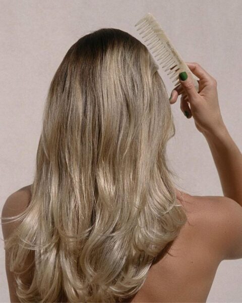 Zdjęcie skupia się na plecach i włosach kobiety, która odwrócona jest tyłem do obiektywu. Ma długie, blond włosy z ciemniejszym odrostem, ułożone w delikatne fale i warstwy. Jej prawe ramię i część pleców są odsłonięte. Trzyma w dłoni o paznokciach pomalowanych na zielono, jasnobeżowy grzebień o szeroko rozstawionych zębach, przeczesując nim włosy. Tło jest gładkie i jasnobeżowe.
