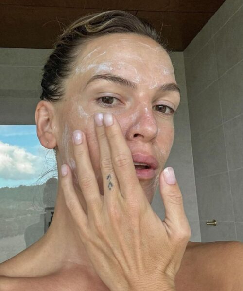 Zbliżenie na twarz kobiety, która nakłada białą, kremową maseczkę lub produkt nawilżający na policzek. Ma ona naturalny makijaż, jasny manicure i tatuaż z dwoma trójkątami na palcu. W tle widać nowoczesną łazienkę oraz fragment widoku na błękitne niebo z chmurami i zielonymi wzgórzami. Scena ilustruje krok z rutyny pielęgnacyjnej z użyciem produktów zawierających składniki aktywne, co jest tematem często poruszanym w przewodniku po składnikach aktywnych dla domowego użytku.