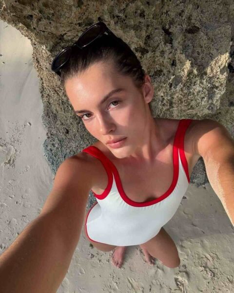 Selfie młodej kobiety na plaży w białym kostiumie kąpielowym z czerwonymi ramiączkami, stojącej na piasku przy skalistym podłożu, z wyraźną miodową opalenizną na skórze.