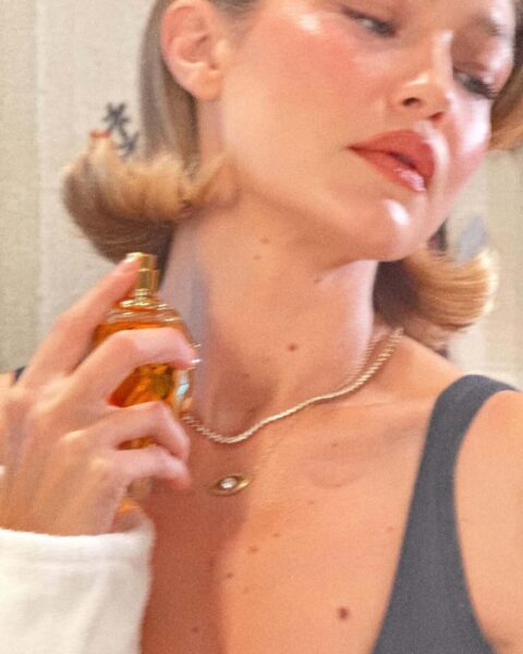 Kobieta o miękko podwiniętych, krótkich włosach trzyma w dłoni złoty flakon perfum pachnących watą cukrową i delikatnie spryskuje nimi szyję. Jej skóra lśni w ciepłym świetle, a subtelny makijaż i złoty naszyjnik dodają całości elegancji oraz kobiecego uroku.