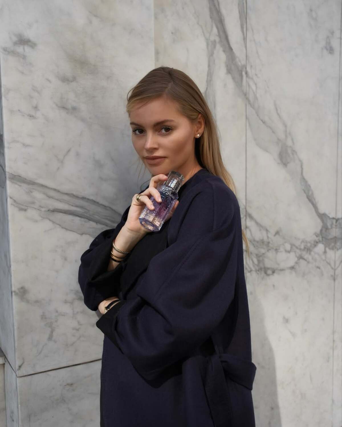 Przytulne perfumy na jesień 2025. Młoda kobieta o jasnych włosach, ubrana w ciemnogranatowy płaszcz z szerokimi rękawami, pozuje opierając się o ścianę z białego marmuru. Trzyma w dłoniach elegancki flakon fioletowych perfum. Ma delikatny makijaż, podkreślony złotymi akcesoriami na nadgarstkach i małym złotym kolczykiem w uchu. Flakon, który obejmuje, symbolizuje idealne przytulne perfumy, otulające ciepłym zapachem.