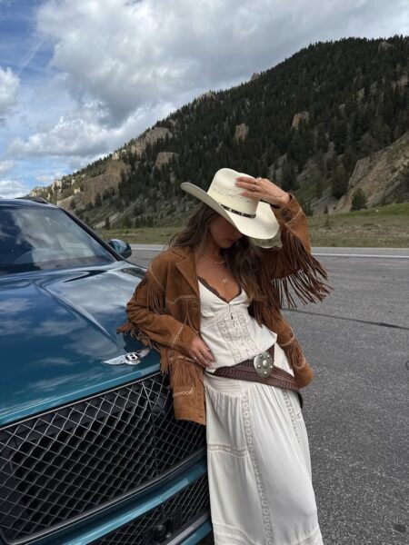 Kobieta w kowbojskim kapeluszu i zamszowej kurtce z frędzlami opiera się o samochód na tle górskiego krajobrazu, stylizacja w klimacie western na jesień 2025.