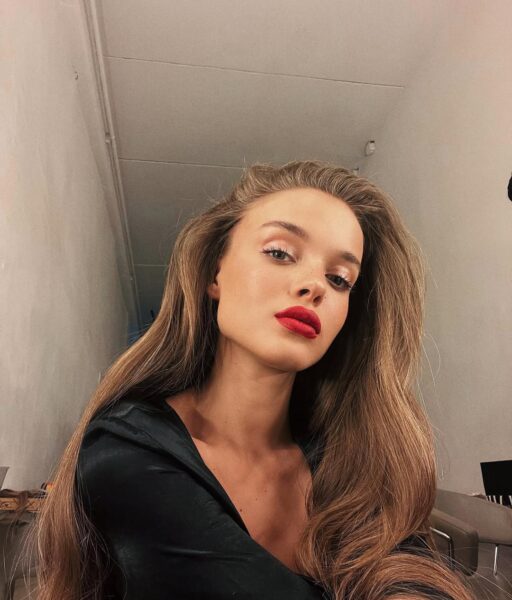 Selfie modelki z długimi, rozpuszczonymi, jasnobrązowymi włosami, opadającymi swobodnie na ramiona. Ma na sobie czarną, satynową sukienkę z głębokim dekoltem. Makijaż jest naturalny z wyjątkiem ust. Wyrazista, matowa czerwona szminka nadaje jej uwodzicielski, retro wygląd. Zdjęcie zostało wykonane w pomieszczeniu z długimi, jasnymi ścianami.