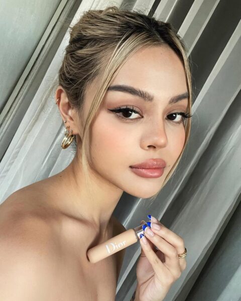 Portret kobiety z upiętymi włosami, trzymającej w dłoni buteleczkę kosmetyku do makijażu marki Dior. Ma na sobie pełny makijaż, w tym eyeliner i naturalne usta. Zdjęcie może być użyte w kontekście makijażu, a także w poradach dotyczących usuwania plam z makijażu z ubrań.