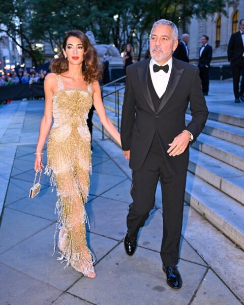 Zdjęcie Amal i George'a Clooneyów, wychodzących z imprezy plenerowej, prawdopodobnie gali. George Clooney ubrany jest w nienaganny smoking z muszką. Amal, trzymająca go za rękę, ma na sobie spektakularną, długą, złotą suknię ozdobioną błyszczącymi frędzlami, z pasującą torebką i wyraźną czerwoną szminką. Ich wyrafinowany, klasyczny wygląd ugruntowuje ich pozycję jako jednej z najbardziej stylowych par w historii Hollywood i współczesnej elity.