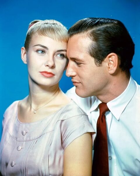 Kolorowe, studyjne ujęcie Joanne Woodward i Paula Newmana na niebieskim tle. Newman w białej koszuli z rozpiętym kołnierzykiem i czerwonym krawatem, pochyla się, dotykając policzkiem czoła Woodward. Woodward ma na sobie jasnoróżową bluzkę z guzikami i delikatny naszyjnik, patrząc lekko w bok. Ta intymność uchwycona na zdjęciu pokazuje, dlaczego Newman i Woodward są często wymieniani jako najbardziej stylowe pary w historii Hollywood i symbol wiecznej elegancji.