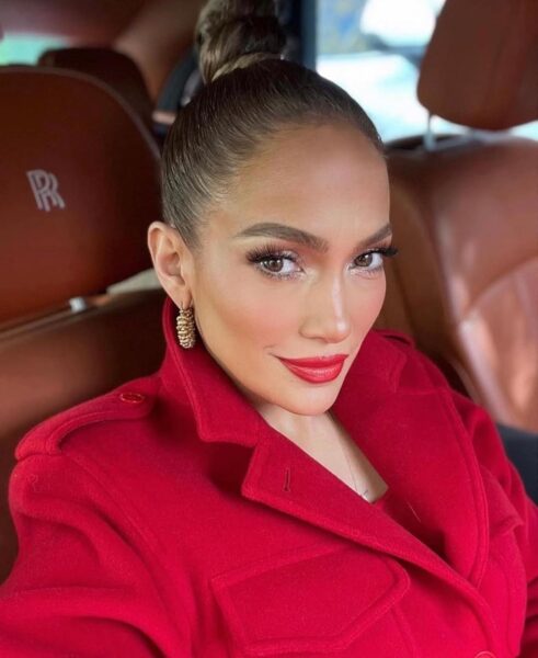 Selfie Jennifer Lopez w samochodzie z brązową, skórzaną tapicerką Rolls-Royce'a, co widać po logo na zagłówku. Ubrana jest w intensywnie czerwoną, wełnianą marynarkę lub płaszcz, a włosy ma spięte w ciasny kok. Jej makijaż jest podkreślony mocnym akcentem na ustach. Złote, teksturowane kolczyki i wyrazisty kolor ust doskonale pasują do jej stroju. Intensywny odcień czerwonej szminki na jej wargach idealnie komponuje się z czerwoną kurtką, tworząc monochromatyczny, ale wyrazisty look.