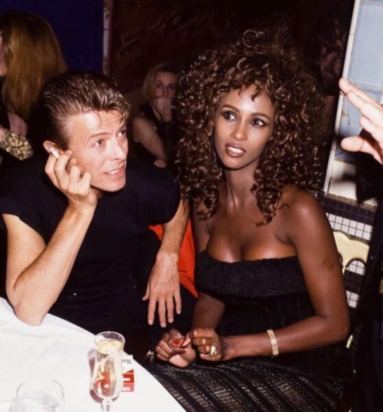 Kolorowe zdjęcie Davida Bowiego i Iman siedzących przy stole. Bowie, ubrany na czarno, z papierosem w dłoni, opiera głowę na drugiej dłoni i patrzy na Iman. Iman, w czarnej sukni bez ramiączek, z burzą kręconych włosów i wyrazistą biżuterią, spogląda w prawą stronę. Ich awangardowy styl na zawsze wpisał ich do kanonu jako jedną z najbardziej stylowych par w historii Hollywood i światowej popkultury.