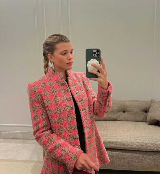 Fashion girl w kultowym, tweedowym żakiecie w odcieniach fuksji i beżu. Stoi w luksusowym, minimalistycznym wnętrzu. Elementy garderoby, które budują status fashion girl: akcesoria nawiązujące do marki (kwiat kamelii) oraz eleganckie, diamentowe kolczyki, które są symbolem wyrafinowanego stylu.
