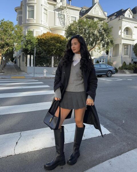 Młoda kobieta w czarnym, długim płaszczu i szarym, plisowanym, krótkim fasonie - idealna spódnica na zimę. Pod spodem ma biały golf i szary sweter w serek. Do tego czarne, wysokie kozaki i czarną torebkę. Stoi na przejściu dla pieszych, a w tle widać jasne kamienice.