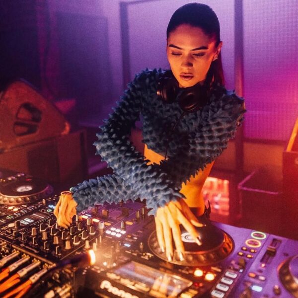 Kim Turnbull jako DJka, skupiona na miksowaniu muzyki na konsoli Pioneer w klubie nocnym. Ma na sobie niebieski, krótki top z długimi rękawami o teksturowanym, wypukłym wzorze. Na szyi wiszą jej czarne słuchawki. Scena jest oświetlona fioletowo-pomarańczowym, nastrojowym światłem.