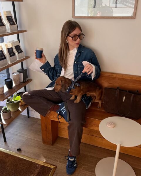 Kobieta siedzi na drewnianej ławce w przytulnej kawiarni, z nogą założoną na nogę, trzymając kubek z kawą i małego jamnika. Ma na sobie jasnobrązowe spodnie, nawiązujące do trendu czekoladowych jeansów na jesień 2025, oraz długą, białą koszulę, na którą luźno narzuciła kurtkę jeansową. Stylowe, ciemne okulary przeciwsłoneczne i dyskretna złota biżuteria dodają jej miejskiej elegancji, a granatowo-brązowe sneakersy podkreślają casualowy charakter ubioru. Obok niej leży duża, zamszowa torba w czekoladowym kolorze, a tło wypełniają półki z kawą i naturalne, drewniane elementy.