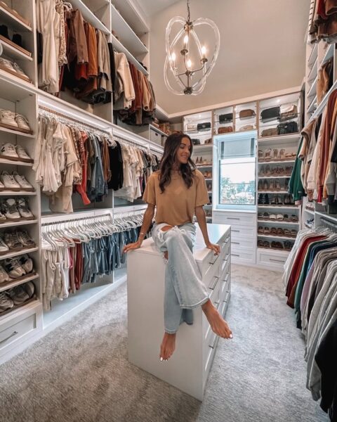 Młoda kobieta w beżowym T-shircie i przetartych jeansach siedzi boso na białej wyspie, znajdującej się pośrodku luksusowej garderoby (walk-in closet). Półki i wieszaki są wypełnione setkami ubrań, butów i torebek, co pokazuje skalę organizacji, którą ułatwia najlepsza aplikacja do organizacji garderoby. W tle widać designerski żyrandol i okno.