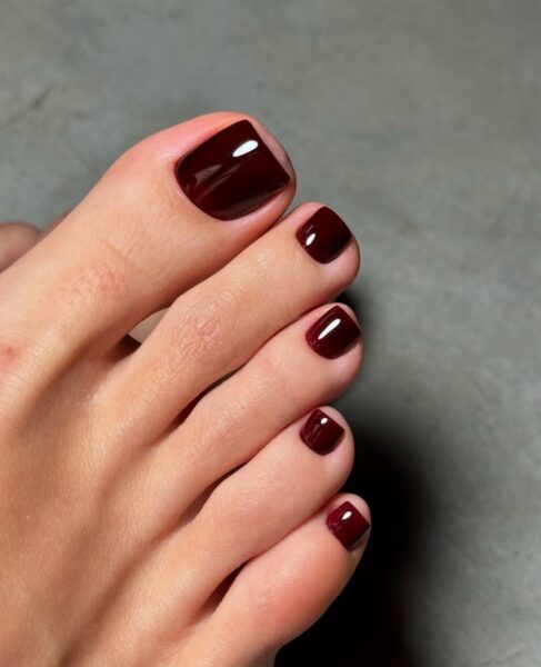 Zbiliżenie na stopę z zadbanym, lśniącym pedicure. Paznokcie u stóp są pomalowane na głęboki, ciemny, bordowy kolor. Ten elegancki kolor jest doskonałą inspiracją na bordowy pedicure na jesień 2025.