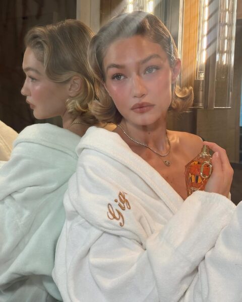 Elegancka kobieta w białym, puszystym szlafroku z haftowanym imieniem na klapie stoi przed lustrem i trzyma w dłoni butelkę perfum pachnących watą cukrową. Jej spokojne spojrzenie, delikatny róż na policzkach i ciepłe oświetlenie tworzą intymną, luksusową atmosferę porannego rytuału piękna.