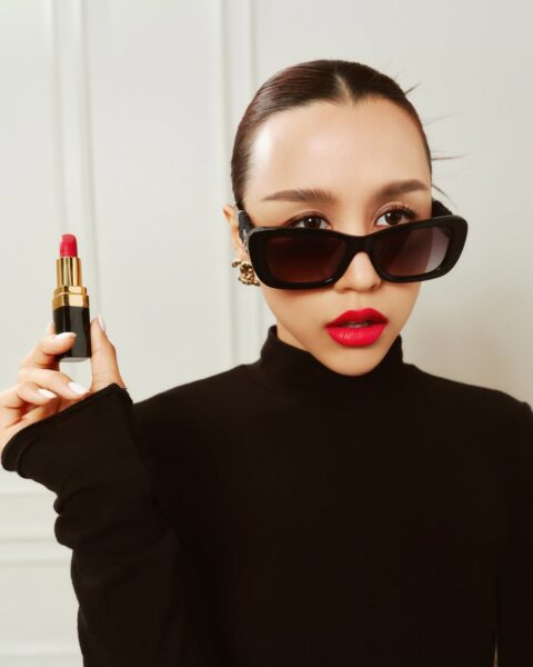 Kobieta w czarnym golfie i dużych, prostokątnych okularach przeciwsłonecznych, trzyma w prawej dłoni elegancką, czarno-złotą szminkę z wyraźnie wysuniętym, intensywnie czerwonym sztyftem. Jej paznokcie są pomalowane na biało. Ma starannie upięte włosy i wargi pomalowane na ten sam żywy odcień, co szminka, którą reklamuje. Na ustach ma już nałożoną czerwoną szminkę, tworząc kontrast z czarnym strojem i okularami, oraz złotym kolczykiem z logo w kształcie łańcucha.