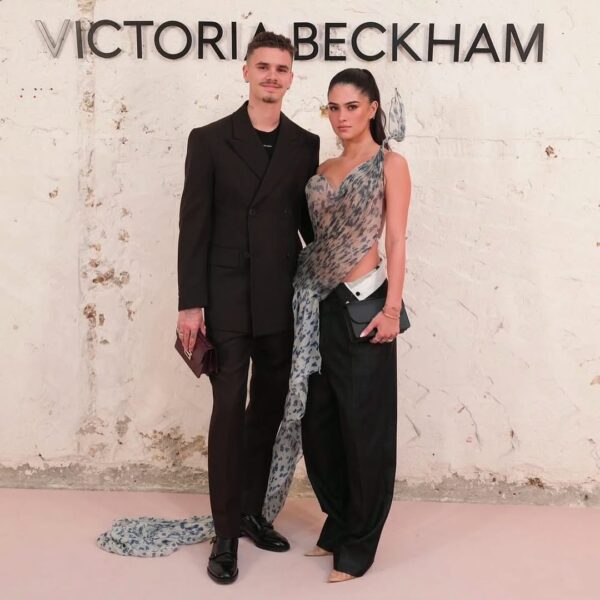 Kim Turnbull i Romeo Beckham na tle ściany z napisem "VICTORIA BECKHAM". Kim ma na sobie asymetryczny top z prześwitującej, wzorzystej tkaniny, która układa się w tren, oraz szerokie, ciemne spodnie. Włosy ma gładko zaczesane do tyłu. Mężczyzna ma na sobie ciemny garnitur z dwurzędową marynarką. Oboje stoją na różowym dywanie.