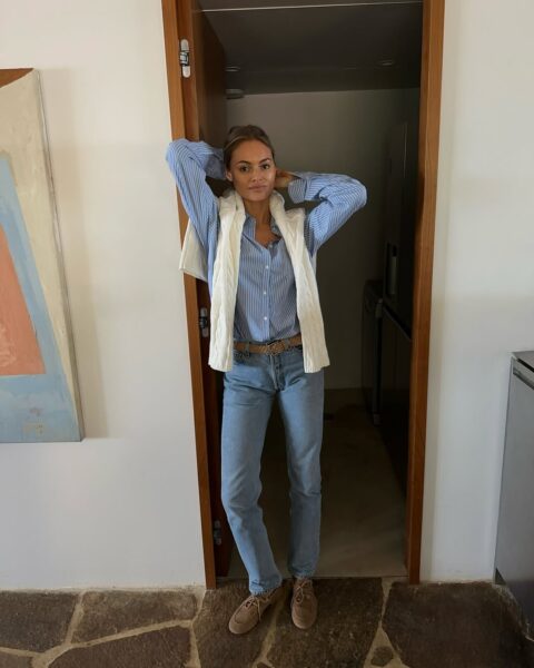 Młoda kobieta w swobodnej, warstwowej stylizacji pozuje, stojąc w framudze drzwi. Ma na sobie jasnoniebieską koszulę w prążki, narzuconą na nią białą, plecioną kamizelkę oraz klasyczne, jasne jeansy z paskiem. Cały zestaw utrzymany jest w stylu preppy/casual i uzupełniają go zamszowe mokasyny lub sznurowane półbuty.