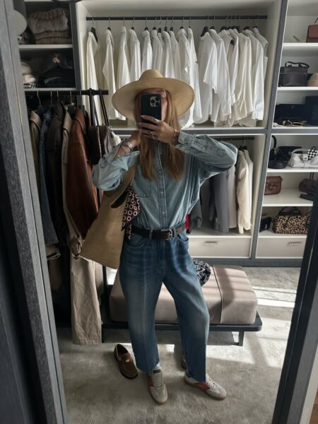 Kobieta w kapeluszu słomkowym i z torebką typu shopper robi sobie selfie w dużym lustrze w swojej przestronnej, uporządkowanej garderobie. Ma na sobie koszulę dżinsową włożoną w luźne dżinsy z paskiem oraz sportowe sneakersy, tworząc wygodną i casualową stylizację. W tle widać rzędy wieszaków z ubraniami, w tym sekcję z samymi białymi koszulami.