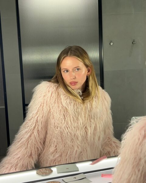 Kobieta w różowym futrzanym swetrze przed lustrem — przykład błędnych połączeń w codziennej rutynie pielęgnacyjnej; jak nie łączyć retinolu z kwasami i witaminą C.