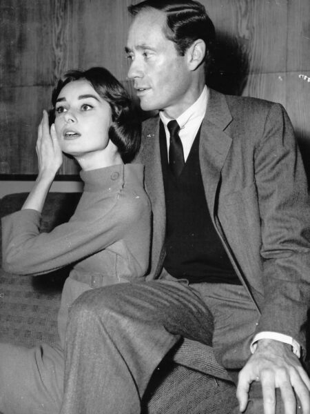 Czarno-białe zdjęcie legendarnej Audrey Hepburn i Mela Ferrera, siedzących blisko siebie. Audrey, w golfie z długim rękawem, z dłonią przy uchu, ma zamyślony wyraz twarzy, spoglądając w lewo. Mel Ferrer w klasycznym garniturze, krawacie i swetrze w serek patrzy przed siebie. Doskonały przykład na to, dlaczego są oni często uznawani za najbardziej stylowe pary w historii Hollywood.