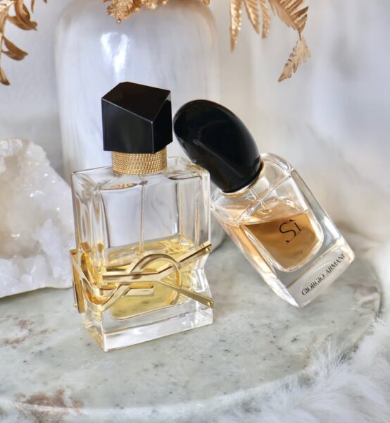 Kompozycja przedstawiająca dwa popularne flakony perfum: YSL Libre oraz Giorgio Armani Sì, ustawione na marmurowej tacy. Tło jest jasne, z widocznymi elementami dekoracyjnymi, takimi jak biała waza, złote liście i kryształ kwarcowy, co nadaje ujęciu luksusowy charakter. Flakony zawierają ciepłe, bursztynowe płyny, które sugerują bogate i otulające aromaty. Perfumy te doskonale sprawdzają się jako przytulne perfumy na jesień, zapewniające poczucie komfortu i elegancji.