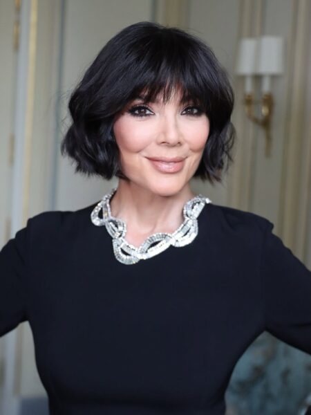 Kris Jenner przed i po. Zdjęcie z bliska przedstawia uśmiechniętą Kris Jenner w czarnej bluzce lub sukience, z krótkimi, ciemnymi włosami z grzywką sięgającą brwi. Uwagę przykuwa duży, masywny naszyjnik z kryształów, ułożony na linii dekoltu. Ma nałożony wyrazisty makijaż. Tło jest jasne i eleganckie, z widocznymi kinkietami.