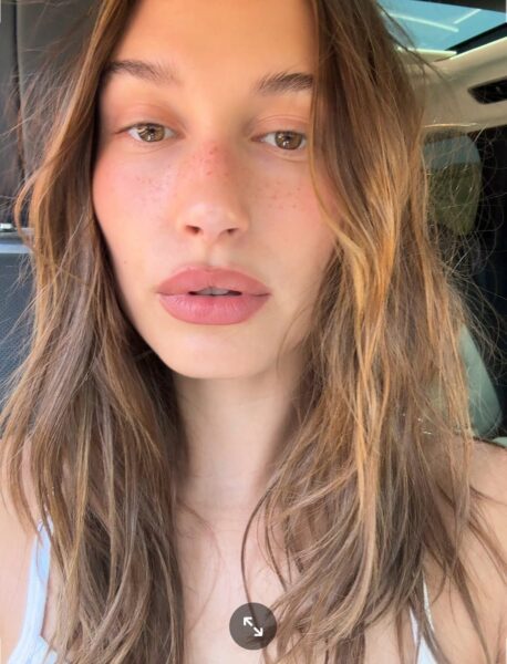 Zdjęcie przedstawia zbliżenie na twarz kobiety, która ma długie, falowane włosy w odcieniach blondu i brązu oraz delikatny, naturalny makijaż z matową, różowo-beżową szminką. Na nosie i policzkach widoczne są piegi. Kobieta ma brązowe oczy i patrzy prosto w kamerę. Ujęcie zrobione jest we wnętrzu samochodu, co sugeruje, że prezentuje szybkie, naturalne urodowe triki z tiktoka lub codzienny, niewymagający makijaż.