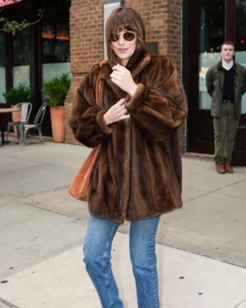 Dakota Johnson idzie po chodniku, na pierwszym planie. Ma na sobie luksusowe, brązowe futro w kolorze czekoladowym, klasyczne dżinsy o prostym kroju oraz stylowe okulary przeciwsłoneczne. Przewieszoną przez ramię ma dużą, zamszową torbę w odcieniu koniaku. W tle widać mężczyznę i ceglaną ścianę budynku.