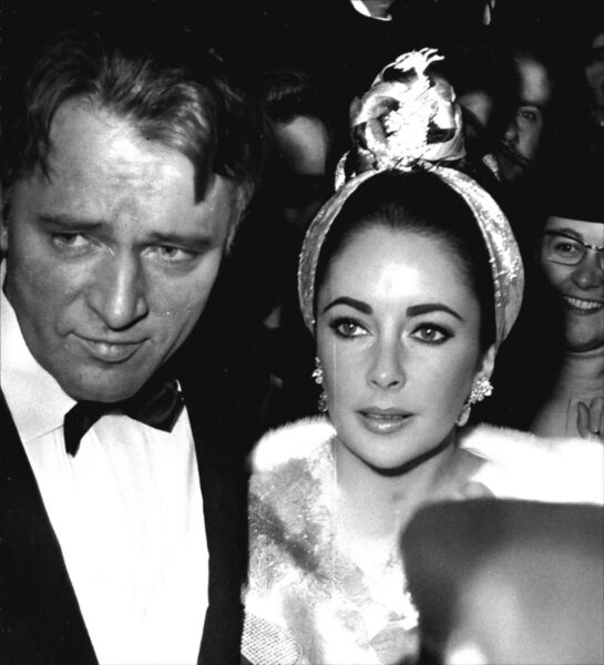 Intrygujące, czarno-białe zbliżenie Elizabeth Taylor i Richarda Burtona na imprezie lub premierze. Taylor ma na sobie efektowne futro i ozdobny turban z błyszczącymi elementami oraz kolczyki, podczas gdy Burton prezentuje klasyczny smoking i muchę. Intensywne spojrzenie Burtona kontrastuje z eleganckim profilem Taylor, co potwierdza, że byli oni jedną z najbardziej stylowych par w historii Hollywood.