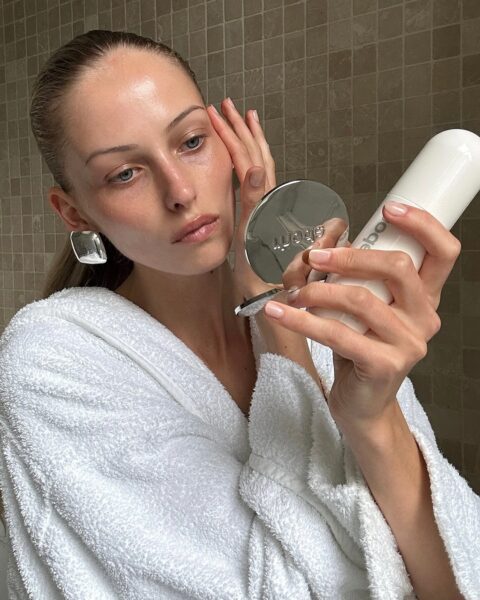 Młoda kobieta w białym szlafroku frotte trzyma w jednej ręce lusterko w metalowej oprawie, a w drugiej białą butelkę, prawdopodobnie serum. Palcami drugiej ręki delikatnie dotyka swojej skóry twarzy, sprawdzając efekt na przykład po nałożeniu serum z niacynamidem. Patrzy intensywnie w lusterko, badając stan swojej cery. Ma duże kwadratowe kolczyki. Tło stanowi łazienka wyłożona beżowymi płytkami.