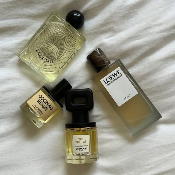 Cztery luksusowe flakony perfum niszowych marek, takich jak Loewe, D.S. & Durga ("Cognac Reign") i Ormaie ("Toi Toi Toi"), leżą na białej, pogniecionej tkaninie. Różnorodność nut zapachowych, na które wskazują nazwy (np. skórzane i koniakowe), jest zgodna z trendami zapachowymi 2025 skupiającymi się na bogatych, złożonych akordach. Zdjęcie to jest wizualną ilustracją świadomego wyboru perfum i eksploracji unikalnych kompozycji.