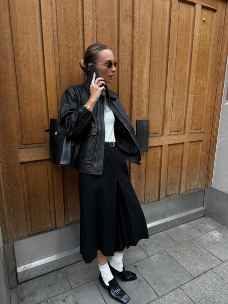 Kobieta rozmawiająca przez telefon, ubrana w minimalistyczny czarno-biały strój: czarną, skórzaną kurtkę, biały top i czarną spódnicę midi z rozcięciem. Połączenie klasycznych elementów i prostych kolorów to doskonały przykład minimalizmu w praktyce w modzie codziennej.
