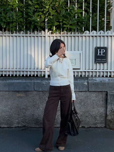 "Kobieta z ciemnymi włosami stojąca przed białą, metalową bramą i szarym murem. Ubrana jest w kremowy, zapinany sweterek, brązowe, szerokie spodnie i beżowe buty. Na szyi ma kilka warstw złotych naszyjników, a na palcu duży pierścionek. Trzyma w lewej dłoni dużą, czarną torebkę. Prawą ręką zasłania część twarzy. Na bramie widoczny jest czarny szyld z logo 'HP HOTEL PARTICULIER' i informacją o usługach fryzjerskich.