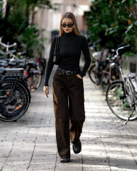 Młoda kobieta o długich blond włosach w ciemnych okularach przeciwsłonecznych, czarnym golfie z długim rękawem, brązowych szerokich dżinsach lub spodniach sztruksowych, czarnym pasku i czarnych butach, idąca brukowaną ścieżką. Ma na ramieniu małą czarną torebkę. Po obu stronach ścieżki widać zaparkowane rowery i zieleń.