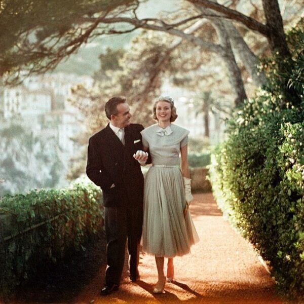 Kolorowe zdjęcie Grace Kelly i księcia Rainiera III, idących ścieżką w ogrodzie. Kelly w eleganckiej szarej sukience midi z bufkami i białymi rękawiczkami, trzyma księcia za rękę. Rainier III, w ciemnym garniturze, uśmiecha się do niej. Ujęcie to, emanujące królewską i hollywoodzką elegancją, podkreśla status tej pary jako jednej z najbardziej stylowych par w historii Hollywood i na świecie.