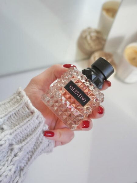 Zbliżenie na dłoń w beżowym, wełnianym swetrze i z czerwonym manikiurem, trzymającą flakon perfum Valentino Donna Born in Roma. Charakterystyczny, pikowany flakon odzwierciedla trend luksusowych zapachów, a posiadanie go jest częścią świadomego wyboru perfum i podążania za globalnymi trendami zapachowymi. Tło jest jasne i rozmyte.