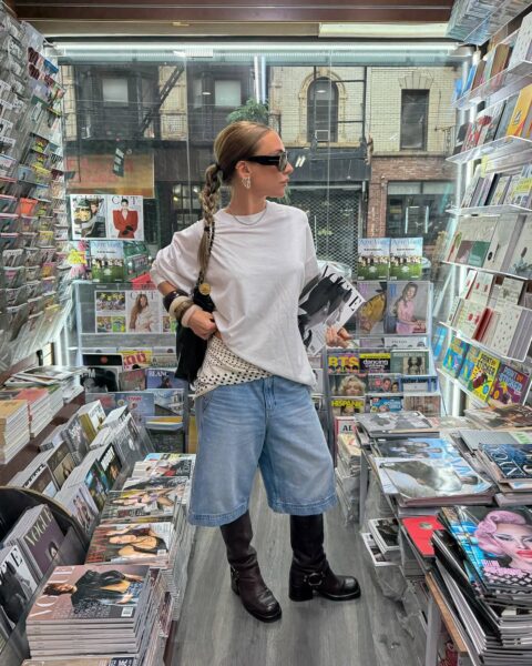 Kobieta w modnym, luźnym stroju stoi w ciasnym wnętrzu kiosku z prasą i magazynami. Ma na sobie szeroką, oversize'ową białą koszulkę, bermudy z jasnego denimu oraz wysokie, skórzane kozaki z klamrami, co jest inspiracją na stroje. Całość dopełniają okulary przeciwsłoneczne i torebka na ramię.
