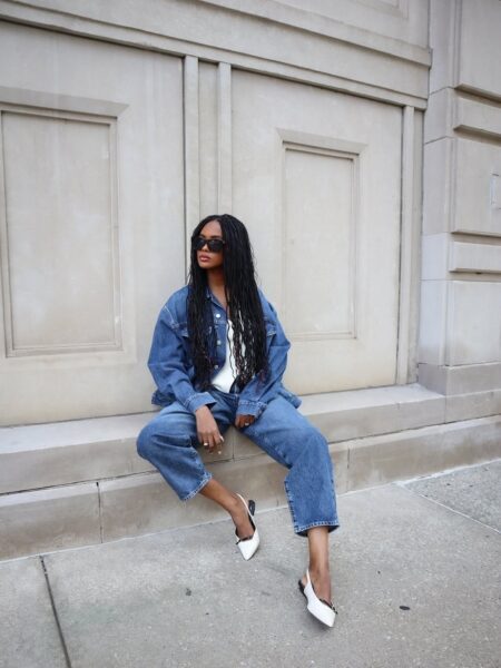 Kobieta w cigarette jeans i oversize'owej kurtce jeansowej siedzi na kamiennym murku. Ma na sobie biały top i czarno-białe czółenka. Długie, ciemne warkoczyki i okulary przeciwsłoneczne uzupełniają jej stylizację.