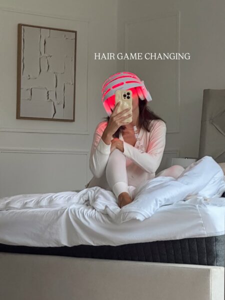 Kobieta siedzi na łóżku, robiąc sobie selfie, a jej twarz jest zasłonięta telefonem. Ma na sobie jasną piżamę i na głowie urządzenie do terapii czerwonym światłem na włosy lub terapia LED na włosy, które emituje intensywne czerwone światło. Na zdjęciu widnieje napis "HAIR GAME CHANGING". W tle widać białą ścianę z dekoracyjnym panelem i abstrakcyjnym obrazem w drewnianej ramie. Otoczenie sypialni jest utrzymane w minimalistycznym, jasnym stylu.