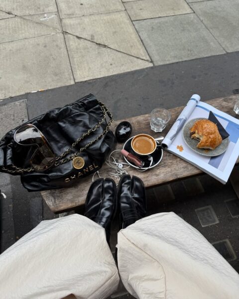 Widok z góry na nogi ubrane w szerokie, jasne spodnie i czarne buty typu Tabi. Osoba siedzi na drewnianej ławce na chodniku, obok niej leży czarna, pikowana torba Chanel, filiżanka espresso, croissant na talerzyku, szklanka wody i słuchawki. Estetyczny zestaw na przerwę kawową.