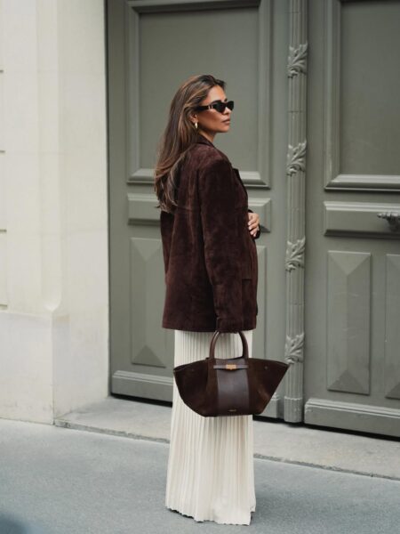 Kobieta w minimalistycznym połączeniu mody jesiennej: brązowej, zamszowej marynarki i długiej, kremowej spódnicy w plisy, trzymająca brązową torebkę. Kolory ziemi i proste formy są idealnym przykładem minimalizmu w praktyce w eleganckim wydaniu.