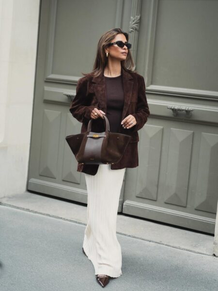Kobieta w brązowej zamszowej marynarce i dopasowanej bluzce, połączonej z długą, prążkowaną spódnicą w kolorze écru. Trzyma brązową torebkę z czarnym akcentem. To stylowa propozycja, jak nosić spódnice zimą.