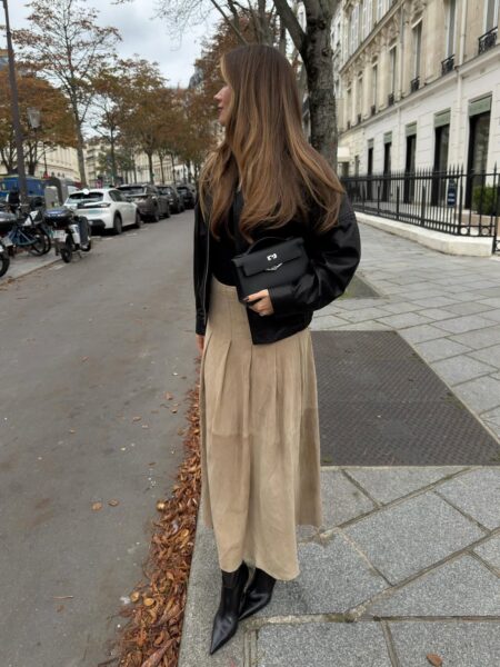 Kobieta stojąca na miejskim chodniku, ubrana w minimalistyczny jesienny strój, łączący czarną skórzaną kurtkę i czarną torebkę ze spódnicą midi w kolorze beżowego zamszu; stylizacja ta, utrzymana w stonowanej palecie barw, to doskonały przykład minimalizmu w praktyce w modzie ulicznej.
