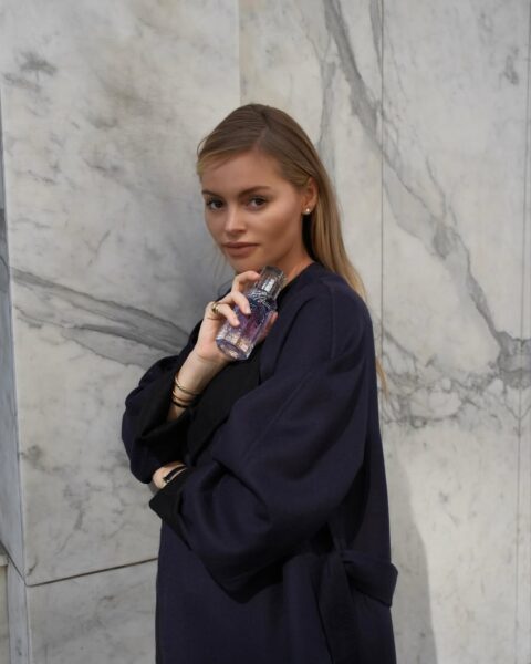 Kobieta w granatowym płaszczu opiera się o marmurową ścianę, z uśmiechem prezentując butelkę perfum pachnących watą cukrową w dłoni.