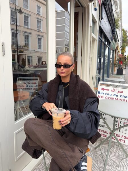 Kobieta siedzi na zewnątrz kawiarni w typowo miejskiej, jesiennej stylizacji, która opiera się na ciemnych kolorach i warstwach. Ma na sobie szerokie czekoladowe jeansy na jesień 2025 i czarny top, a na ramionach zarzuciła puchową, czarną kurtkę w stylu bomber. Całość została urozmaicona ciemnobrązowym swetrem, przewiązanym nonszalancko na ramionach. Kobieta trzyma w rękach kubek z kawą mrożoną, ma okulary przeciwsłoneczne w prostokątnej oprawie, włosy ma związane w gładki kok i nosi sportowe buty.