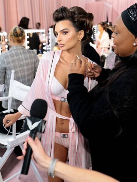 Modelka w bieliźnie i szlafroku w różowo-białe paski jest przygotowywana na backstage'u Victoria's Secret. Makijażystka nakłada jej kosmetyk na szyję i dekolt, a w tle widać mikrofon i inne osoby. Scena doskonale pokazuje, że profesjonalne triki modelek zza kulis obejmują pielęgnację i makijaż nie tylko twarzy, ale i całego ciała.