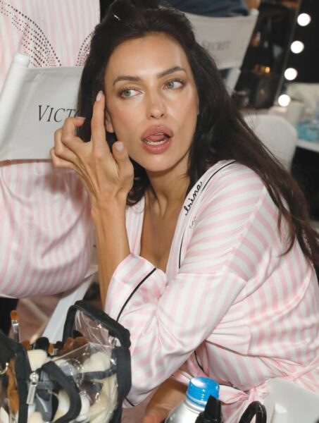 Zdjęcie przedstawiające modelkę w satynowej piżamie w biało-różowe paski, siedzącą na fotelu z logo Victoria's Secret. Modelka w lustrze poprawia sobie delikatnie makijaż, dotykając palcem skóry wokół oka. Ta scena to uchwycenie momentu, w którym w ruch idą ostatnie, kluczowe triki modelek zza kulis przed wielkim pokazem.