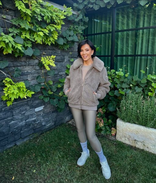 Kris Jenner retusz zdjęć. Zdjęcie przedstawia Kris Jenner pozującą na zewnątrz, w ogrodzie, na tle kamiennego muru porośniętego bluszczem i dużego okna. Ubrana jest w sportowy, monochromatyczny strój w odcieniach beżu i taupe: puszysty, pluszowy (teddy) żakiet/kurtkę zapinaną na zamek, dopasowane błyszczące legginsy i beżową bluzę. Ma na sobie białe buty sportowe, jasnoniebieskie skarpetki z napisem "alo" i małe kolczyki. Jej włosy są krótko ścięte i ciemne. Uśmiecha się, trzymając ręce w kieszeniach.